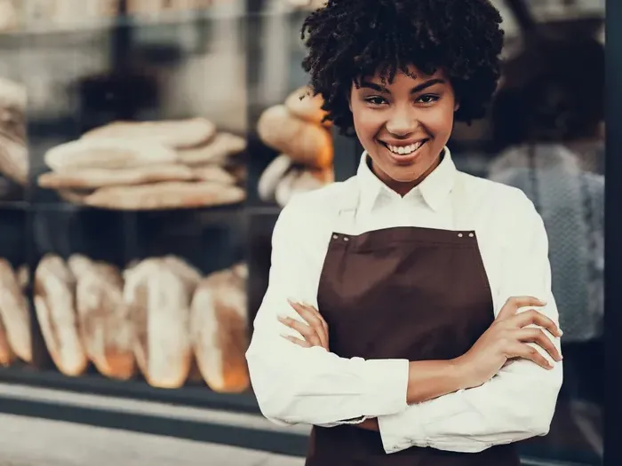 Smiling bakery owner in brown apron with arms crossed, fresh loaves behind window display, welcoming customers